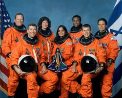 Seven crew members standing in orange pressure suits and holding a larger version of their mission patch.