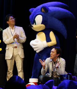 A Sonic the Hedgehog costume performer is flanked by Naka and Oshima in front, while the two men are applauding.