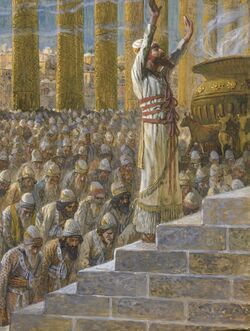 In the foreground, a bearded man dressed in an impressive white robe and head-dress raises his hand to heaven. Behind him, a large crowd bows in prayer.