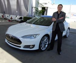 Musks stands, arms crossed and grinning, before a Tesla Model S