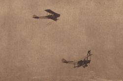 Two biplanes flying, with Ormer Locklear standing on the top wing of the lower airplane