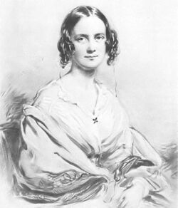 Three-quarter length portrait of woman aged about 30, with dark hair in centre parting straight on top, then falling in curls on each side; she smiles pleasantly and is wearing an open-necked blouse with a large shawl pulled over her arms