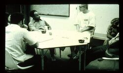 B/w photo of several white and one black person who are sitting at a table with a whiteboard on the wall behind. They are all looking to one white male person sitting at the table wearing a white t-shirt with a number on the chest.