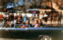 John F. Kennedy is seen sitting in a limousine, waving with a crowd on Elm Street, Dealey Plaza.