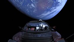 Photograph of the black emptiness of space, with planet Earth partly in shadow in the background. In the foreground is an open-top red convertible sports car, viewed from the front over the hood, with a mannequin in the driving seat that is wearing a white-and-black spacesuit