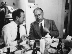 A young man in a white shirt and tie and an older man in suit and tie sit at a table, on which there is a tea pot, plates, cups and saucers and beer bottles.