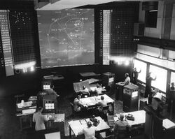 An electronic simulation room at the Naval War College during a 1958 wargame: against the far wall, a large map shows the outline of landmasses and some firing solutions. Suited men sit at desks on the floor, papers in front of them, most staring up at the map. Against the right wall, uniformed ensigns plot ship locations on (washed-out) screens.