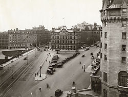 A sepia photograph from the fourth floor of a building, overlooking a triangular public plaza, many old cars with canvas tops are parked in the square. Neo-gothic buildings make up two borders of the square, and a set of tram tracks comprise the third