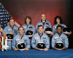 Picture of the seven crew members in flight suits and holding their helmets