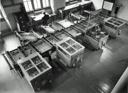 Looking down from above at a room at an intricate mechanical device which fills the room. In the background, a man sits at a desk next to a filing cabinet.[14]