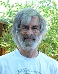 Portrait of a Caucasian man in his seventies with medium-length gray hair and a full gray beard, wearing glasses and a T-shirt.