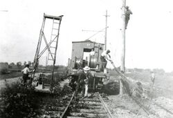 This black and white photograph shows construction workers raising power lines next to the railroad tracks of the Toledo, Port Clinton, Lakeside Railroad tracks in a rural area. The workers are using a railroad car as their vehicle to carry supplies and themselves down the line. It was taken in approximately 1920.