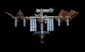 A forward view of the International Space Station with limb of the Earth in the background. In view are the station's sixteen paired maroon-coloured main solar array wings, eight on either side of the station, mounted to a central integrated truss structure. Spaced along the truss are ten white radiators. Mounted to the base of the two rightmost main solar arrays pairs, there are two smaller paired light brown- coloured ISS Roll-out Solar Arrays. Attached to the centre of the truss is a cluster of pressurised modules arranged in an elongated T shape. A set of solar arrays are mounted to the module at the aft end of the cluster.