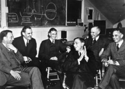 Six men in suits sitting on chairs, smiling and laughing