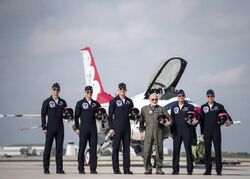 Five men in blue jump suits pose with Aldrin in an olive jump suit on the runway in front of a white F-16
