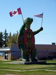 A large turtle statue standing on two legs and holding a Canadian flag in one hand an American flag in the other.