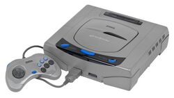 A Japanese Sega Saturn console. It is a gray system that resembles a DVD player. Attached is a gray controller with a dark gray D-pad on the left side and six buttons (three bigger black ones labeled A, B, and C, and three smaller blue ones labeled X, Y, and Z).