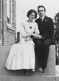 A young man in a suit and tie and a young woman in a light coloured dress sit on a stoop, holding hands