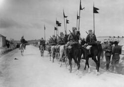 Black-and-white photo of mounted soldiers with middle eastern headwraps, carrying rifles, walking down a road away from the camera