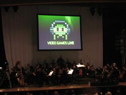A photograph of an orchestra on a dimly lit stage. Above the group is a projection screen with a black, white, and green image of pixel art. The pixel art is an oval object wearing headphones with eyes and four tentacles. Below the pixel art is the phrase "Video Games Live".