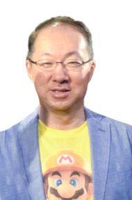 A Japanese man in his 50s. He has short brown hair and glasses and is smiling towards the camera. He is wearing a light blue blazer and a yellow shirt with Mario on it underneath.