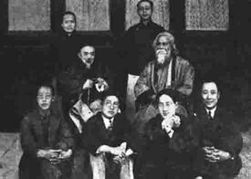Posed group black-and-white photograph of seven Chinese men, possibly academics, in formal wear: two wear European-style suits, the five others wear Chinese traditional dress; four of the seven sit on the floor in the foreground; another sits on a chair behind them at centre-left; two others stand in the background. They surround an eighth man who is robed, bearded, and sitting in a chair placed at centre-left. Four elegant windows are behind them in a line.