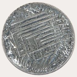 A shiny silver-white medallion with a striated surface, irregular around the outside, with a square spiral-like pattern in the middle.