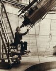 A man sitting on a chair mounted to a moving platform, staring through a large telescope.