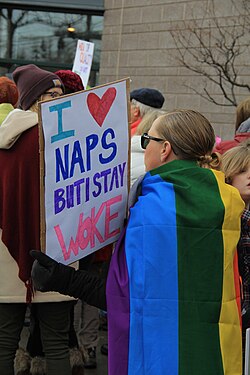 A woman draped in a rainbow flag and wearing sunglasses, standing with her back to the camera and holding a hand-lettered sign reading, "I [heart symbol] Naps But I Stay Woke"