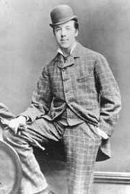 Oscar Wilde posing for a photograph, looking at the camera. He is wearing a checked suit and a bowler hat. His right foot is resting on a knee-high bench, and his right hand, holding gloves, is on it. The left hand is in the pocket.