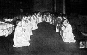 Two rows of little boys, about 20 in total, kneel before their beds in the dormitory of a residential nursery. Their eyes are shut and they are in an attitude of prayer. They wear long white night gowns and behind them are their iron-framed beds.