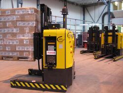 A yellow rectangular wheeled forklift robot in a warehouse, with stacks of boxes visible and additional similar robots visible behind it