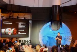 Musk speaks to a crowd of journalists. Behind him is a lighted tunnel.