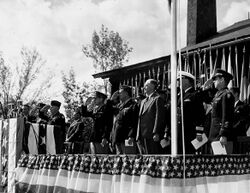 Men in suits and uniforms stand on a dais decorated with bunting and salute.